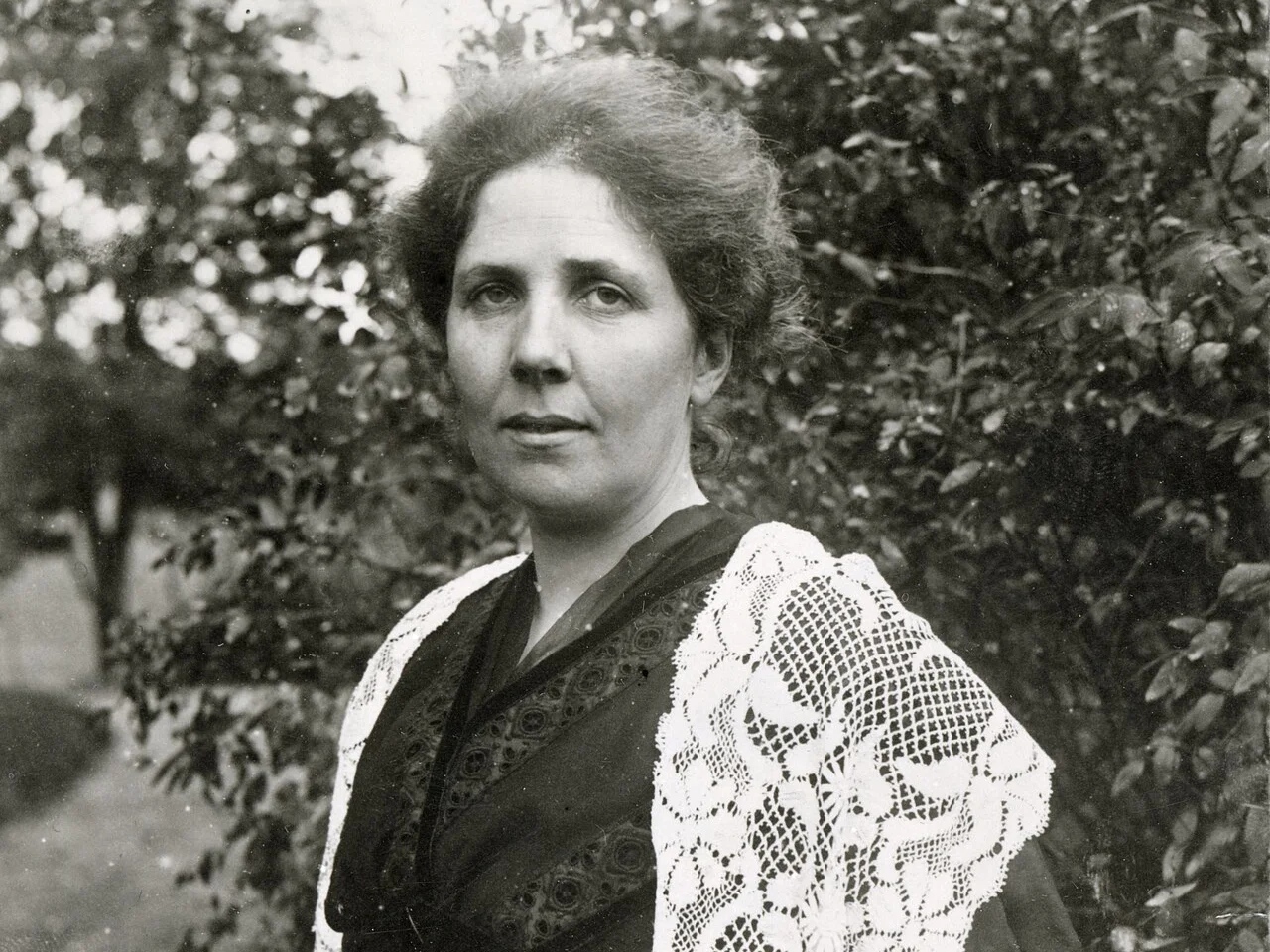 Svartvitt porträtt av Elsa Beskow med uppsatt hår och spetskrage framför gröna buskar